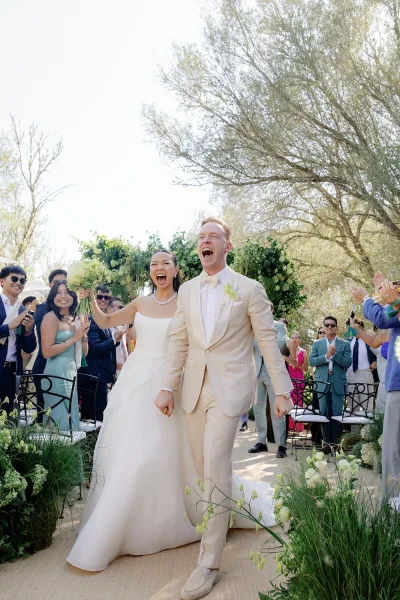 Ceremony recessional with a couple walking down the aisle, the bride lifting her bridal bouquet as guests cheer beneath garden trees and blue sky.