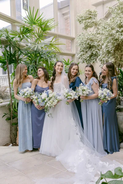 Bridesmaid group photo of bride with bridesmaids in mismatched blue dresses holding bouquets, standing by greenhouse windows and plants with veil accent