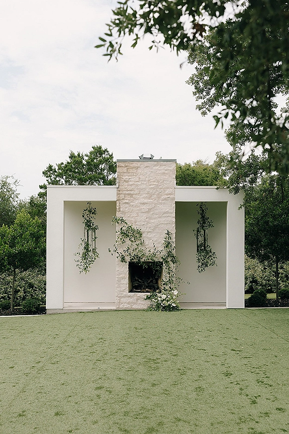 Wedding ceremony backdrop with hanging greenery and vines on a white wall beside a stone fireplace, set on a lawn under trees
