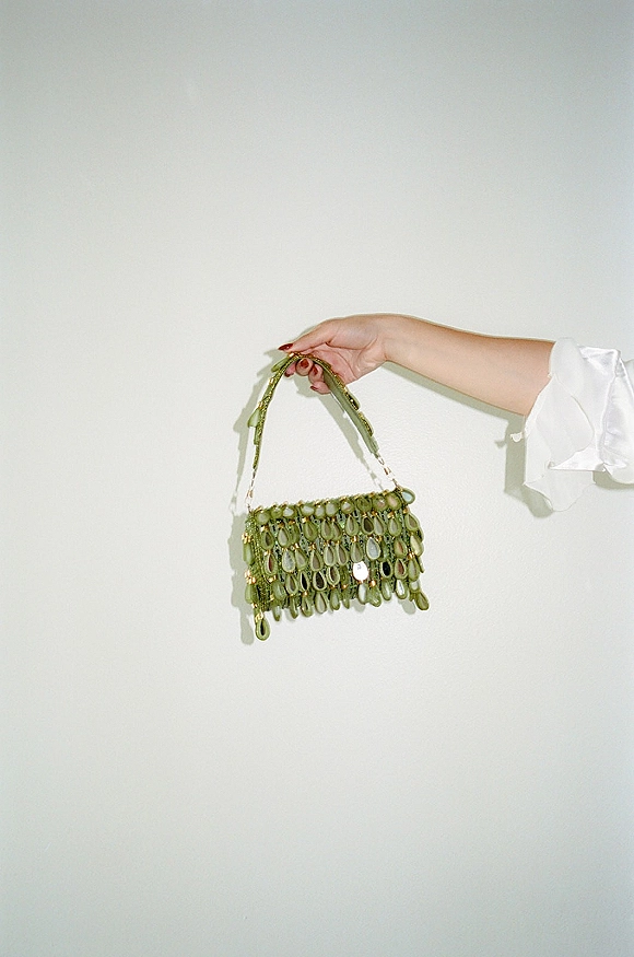 Bridal purse, a green beaded wedding day handbag with gold hardware and handle, held by manicured hand in white satin robe on white wall shadow