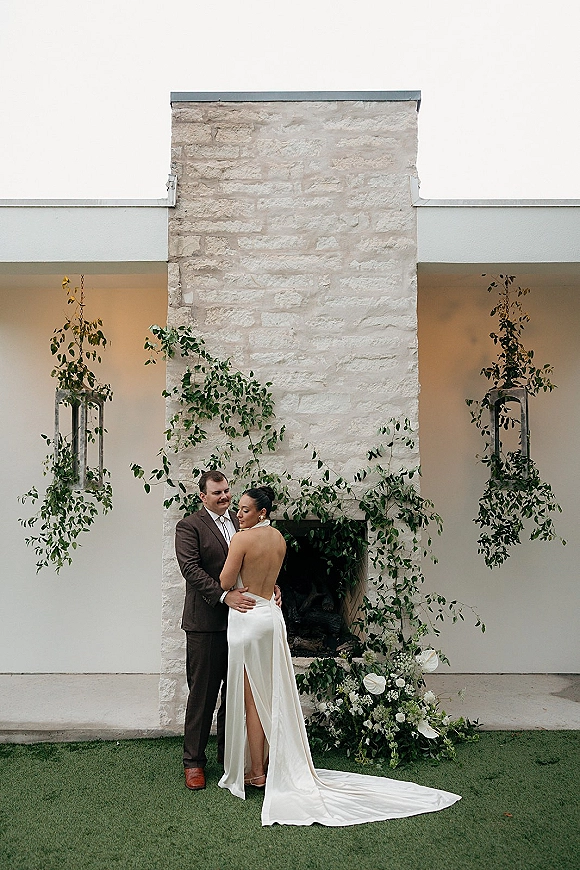 Couple portrait of bride and groom embrace, her backless wedding dress with train beside a stone fireplace with greenery vines and lanterns