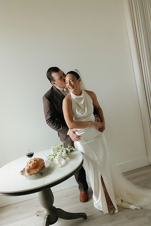 Couple portrait of groom kissing bride’s cheek as she laughs in a halter gown and veil, beside a table with bouquet and wine