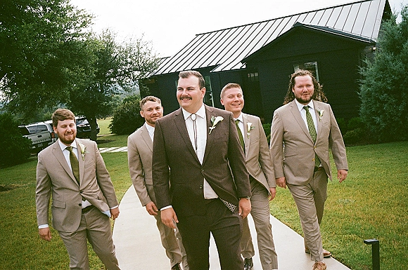 Groomsmen portrait of the groom with groomsmen walking in tan suits and boutonnieres on a concrete walkway by a modern barn venue