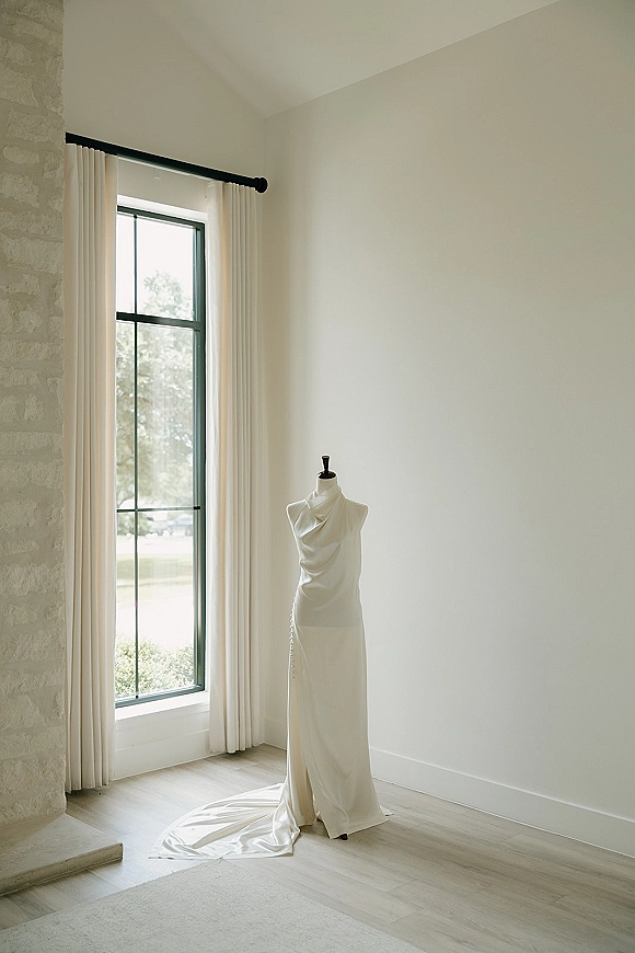 Wedding dress in satin on a mannequin with a draped neckline and chapel train, back buttons glowing in window light beside a stone fireplace