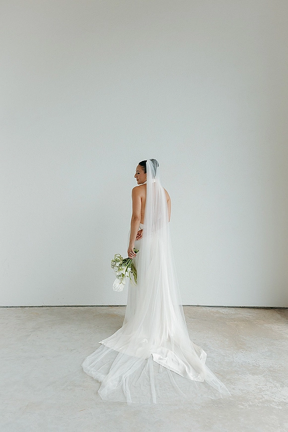 Bridal portrait of a bride looking over her shoulder in a strapless gown with long veil train and bouquet against a white wall studio backdrop