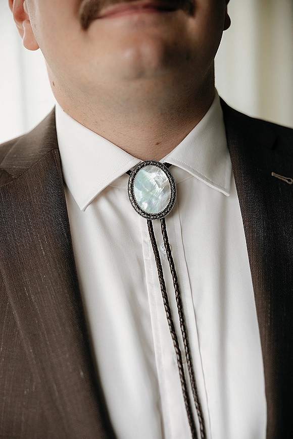 Bolo tie on a groom, featuring an oval stone clasp and braided leather cords over a white dress shirt in soft window light