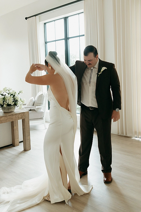 First dance moment as bride and groom dancing by large windows, her veil flowing behind a low back dress in a bright room
