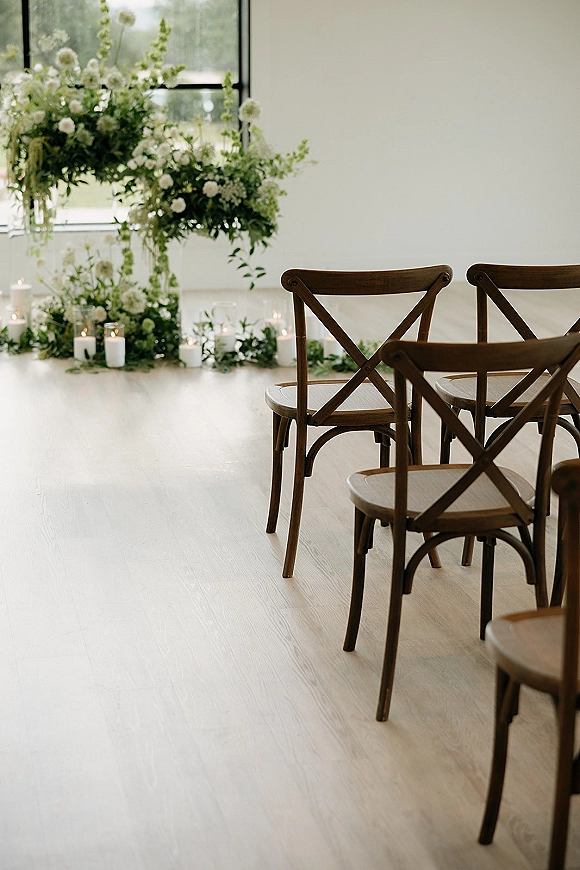 Ceremony aisle decor with a candle lined aisle, greenery garland, and white floral arrangements beside wood crossback chairs in an airy room