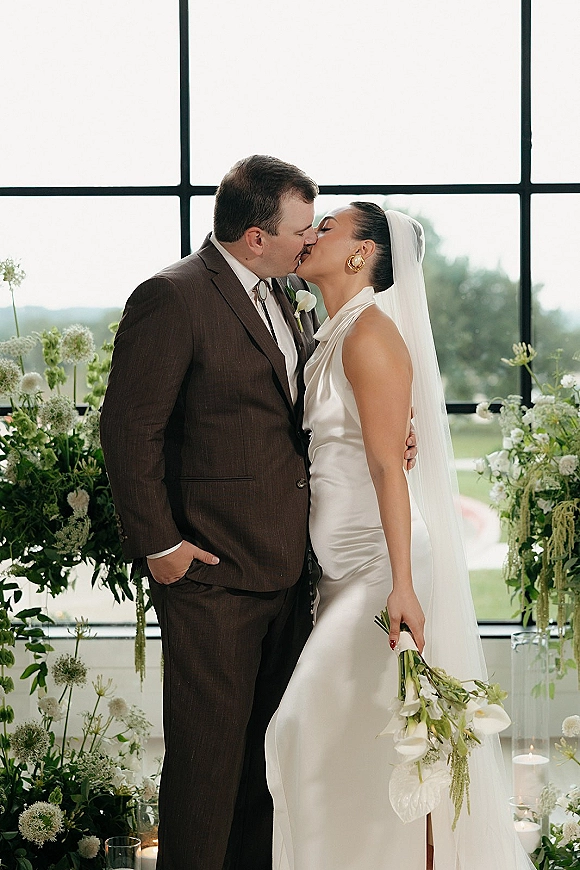 Wedding kiss portrait of bride and groom kissing by a large grid window, her veil and calla lily bouquet lit by soft natural light