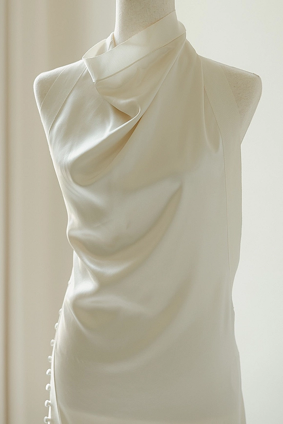 Wedding dress close-up of a satin wedding dress with draped cowl neckline and side buttons on a mannequin in soft window light