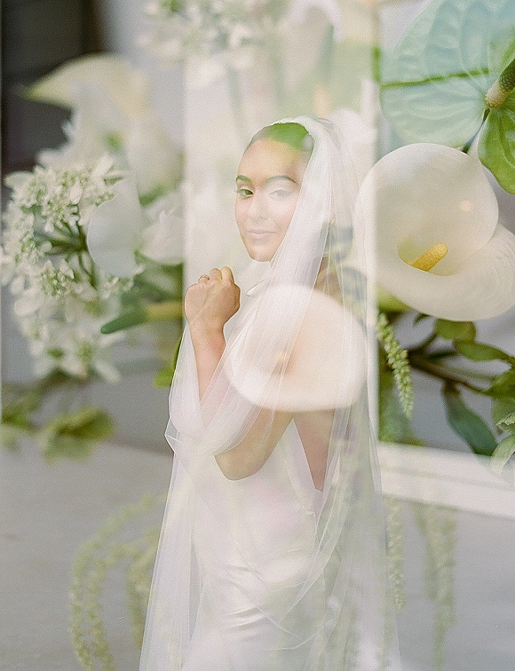 Bridal portrait of a bride with veil in a strapless satin gown, looking over her shoulder in a garden, holding white calla lilies