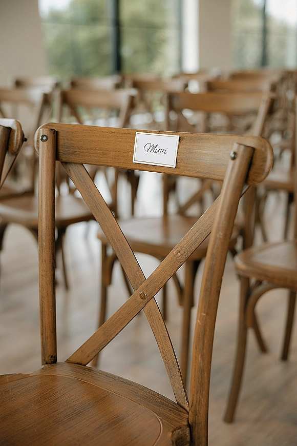 Wedding seating cards clipped to wooden cross back chairs, marking reserved seats in an indoor ceremony room with large windows and greenery outside