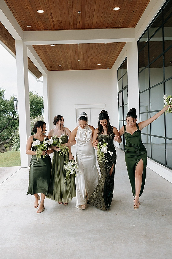 Bride and bridesmaids walking with bride under a covered patio, carrying white calla lily bouquets with greenery, veil flowing behind