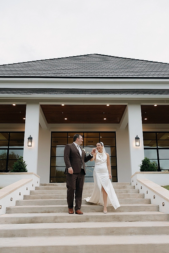 Couple portrait of bride and groom on steps, holding hands as he helps her descend in a veil at a modern entrance with columns