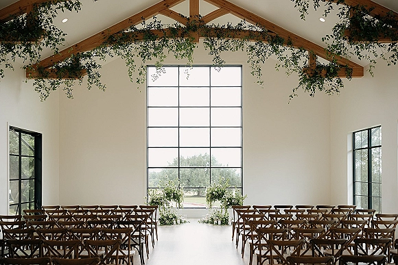 Ceremony setup for an indoor wedding ceremony with wood crossback chairs lining the aisle beneath hanging greenery and candlelit altar florals by large windows