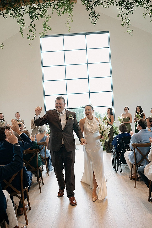 Ceremony recessional as newlyweds walk hand in hand down the aisle, bride in satin halter gown with white bouquet, guests cheering under hanging greenery