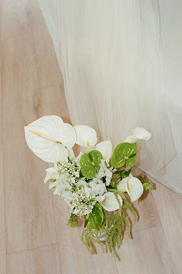 Bridal bouquet with anthurium wedding bouquet blooms and white calla lilies in a glass vase on a wood floor beside sheer drape