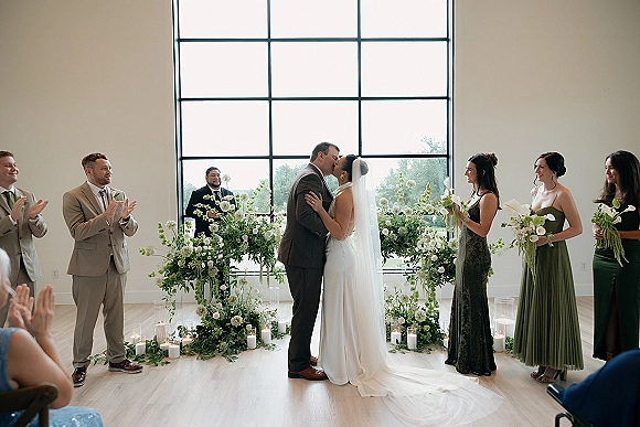 Ceremony kiss as bride and groom kiss at the altar, her long veil flowing, bridesmaids in green with white bouquets, candles below large window