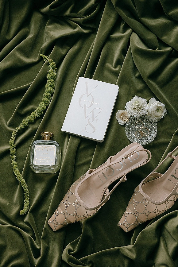 Wedding flat lay with wedding invitation suite, rings in a crystal dish, perfume, bridal shoes, and white flowers on green satin backdrop