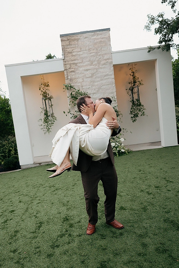 Wedding kiss portrait of groom carrying the bride kiss, her strapless dress and gold earrings against a modern white facade with greenery planters
