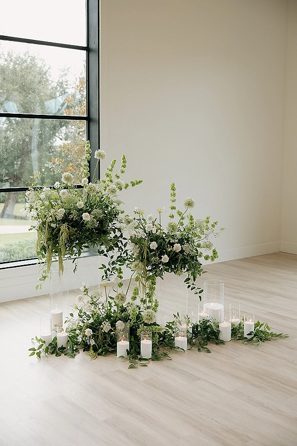 Ceremony altar decor with modern wedding altar accents, white florals and greenery on clear plinths, candles by a black-framed window