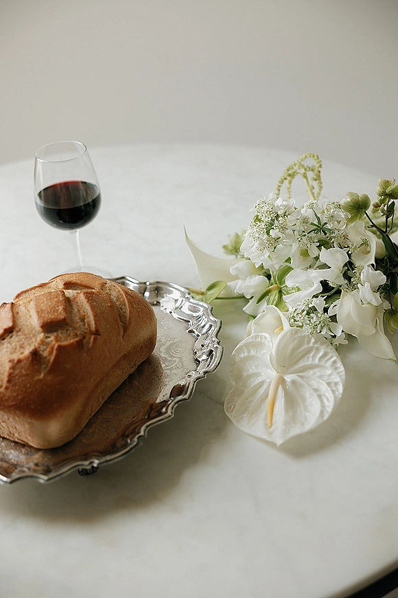 Wedding tablescape with a wedding bread display on a silver serving tray, red wine glass, and white anthurium flowers on a white table