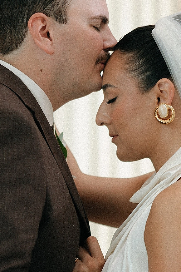 Couple portrait with a groom in a brown suit giving a forehead kiss wedding moment to a bride in a white halter dress against a neutral backdrop