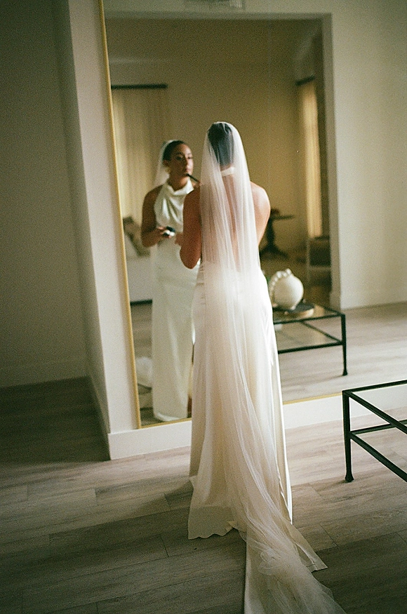 Bridal getting ready bride applying lipstick in a mirror, wearing a sleek wedding dress and long veil in a window-lit room with curtains