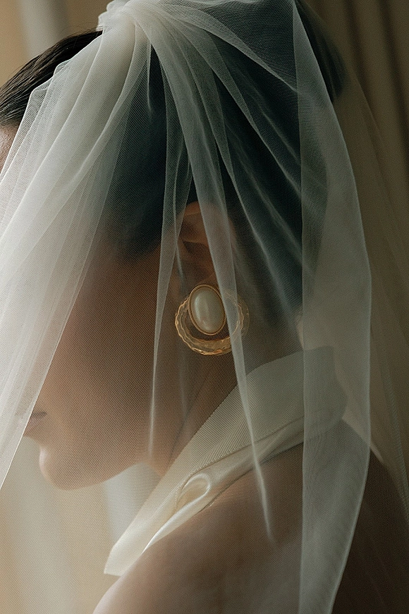 Bridal portrait of a bride with veil in side profile, sheer tulle over her face highlighting a pearl earring against a neutral indoor backdrop