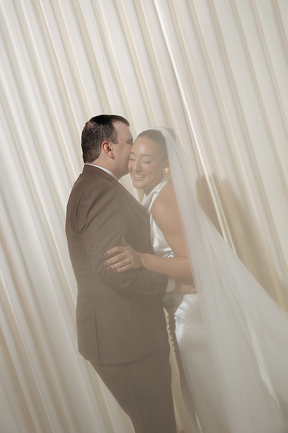 Couple portrait of bride and groom embrace as he kisses her forehead under a sheer veil, soft indoor light with white curtains behind