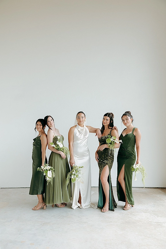 Bride with bridesmaids in green dresses holding calla lily bouquets, bride in white satin gown and veil against a white studio wall