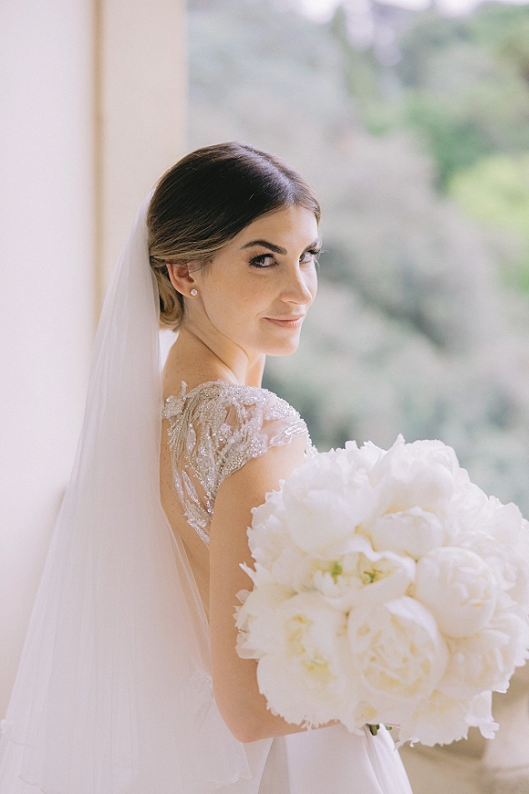 Bridal portrait of a bride holding bouquet, looking back over her shoulder with a veil and white peony bouquet in soft greenery outdoors