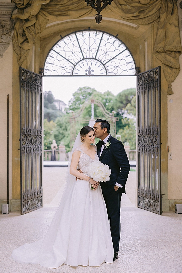 Wedding couple portrait of bride and groom hug under an ornate archway, groom kissing her forehead as she holds a white bouquet