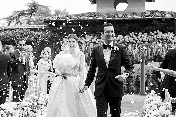 Wedding recessional as newlyweds walking down aisle hand in hand while guests toss flower petals in an outdoor courtyard with floral wall