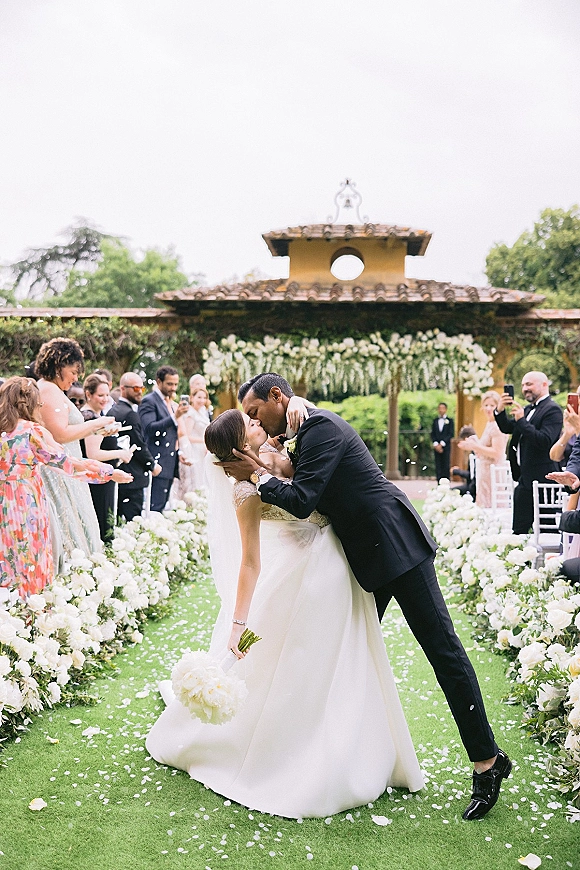 Wedding kiss portrait of newlyweds in a dip, bride in veil with white bouquet as guests cheer along a floral-lined garden aisle