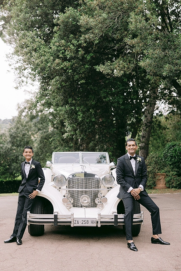 Groom portrait in a black tuxedo leaning on a vintage white car, bow tie and boutonniere visible, on a garden driveway with trees