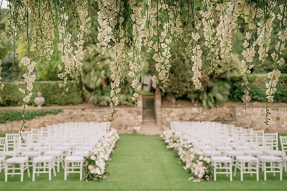 Ceremony aisle decor with an outdoor wedding ceremony setup, white chairs lining a grass aisle with lush white florals and hanging greenery overhead