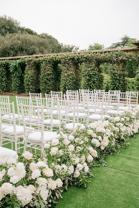 Outdoor ceremony setup with white chiavari chairs lining a grass aisle runner, white rose florals and greenery garland on a garden lawn