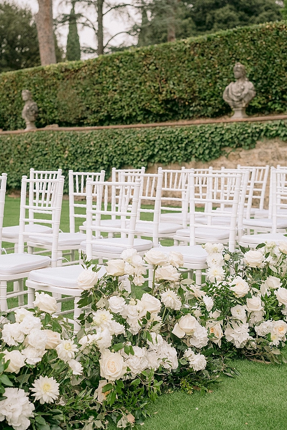 Ceremony aisle decor with white wedding aisle flowers and greenery garland lining a garden lawn, flanked by white Chiavari chairs