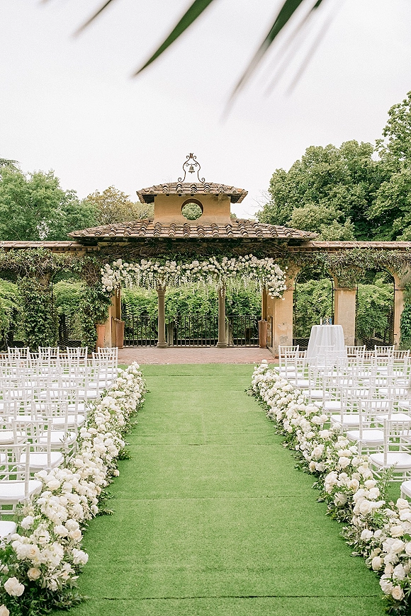 Ceremony setup for an outdoor wedding ceremony with white aisle flowers, a floral arch, and chairs in a garden courtyard pavilion setting