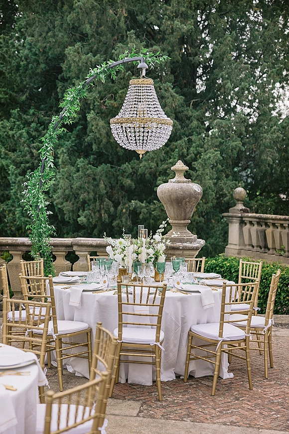Reception tablescape with an outdoor reception table of round white linens, gold Chiavari chairs, taper candles and floral centerpiece under a crystal chandelier on a brick patio
