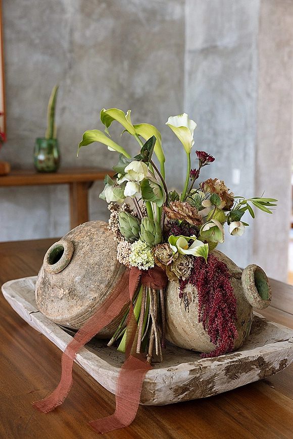 Wedding bouquet with calla lilies and artichokes, hellebores, dried amaranthus, and greenery on a wood table by a concrete wall