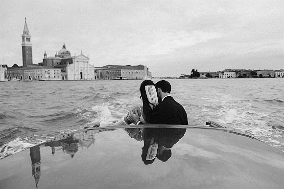 Couple portrait of newlyweds on a motorboat, groom’s arm around bride in long veil, with water and city skyline under cloudy sky