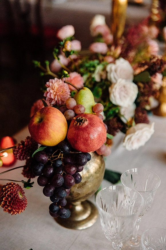 Wedding centerpiece with a fruit wedding centerpiece of pomegranate, grapes, and pears among roses and dahlias in a gold compote bowl with taper candles