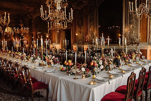 Reception tablescape with brass candelabra centerpiece and taper candles, floral and fruit arrangements on long table in ornate ballroom with chandeliers