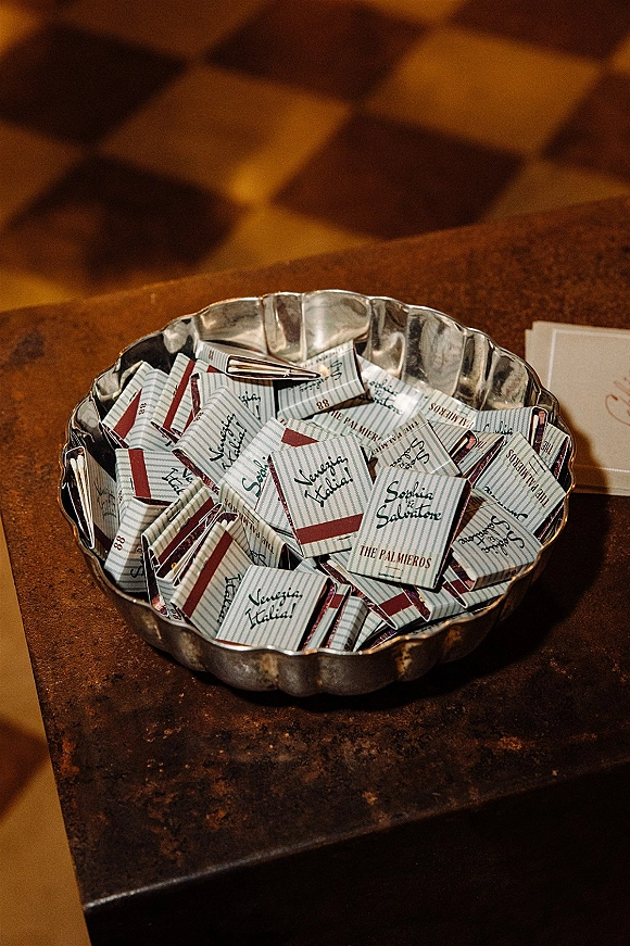 Wedding favors in a metal bowl, featuring personalized matchbox favors with striped labels and printed names on a brown velvet surface
