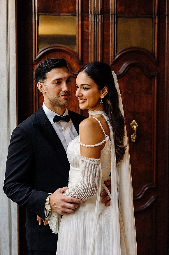 Couple portrait of bride looking over shoulder in pearl strap gown and veil as groom holds her waist by wooden double doors