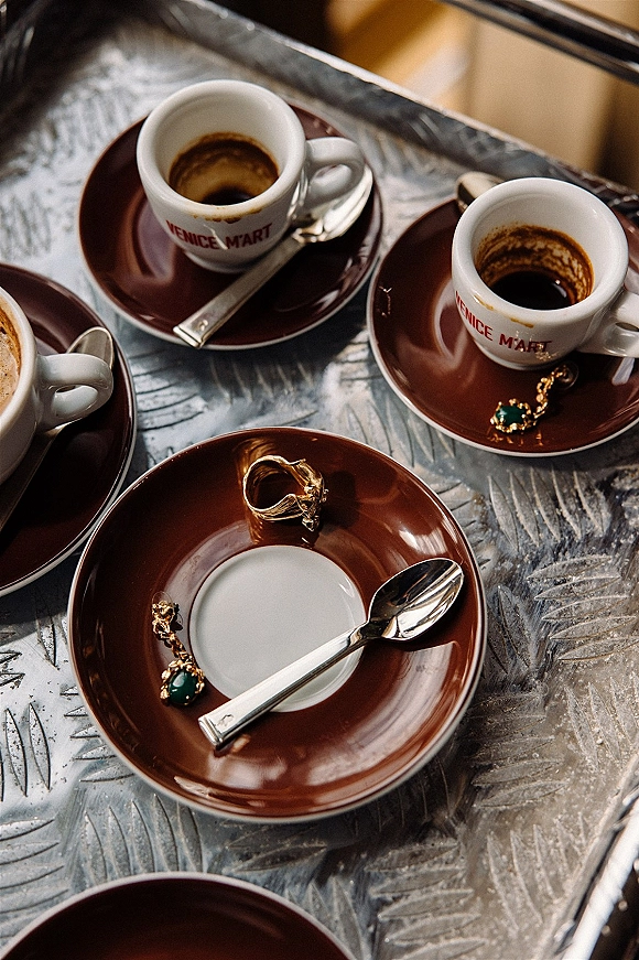 Wedding rings in a wedding ring flatlay beside espresso cups on saucers, with a gold ring and green gemstone bracelet on a metal café table