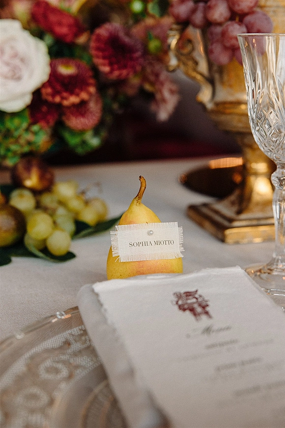 Wedding place card on a pear place card with handwritten name tag tied in frayed ribbon beside a menu with crest and crystal goblet on a reception table