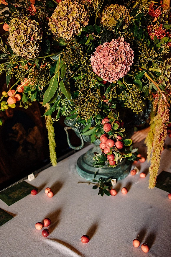 Wedding centerpiece with hydrangea centerpiece blooms and greenery in a metal urn vase, with berry clusters and hanging amaranthus on a dim reception table
